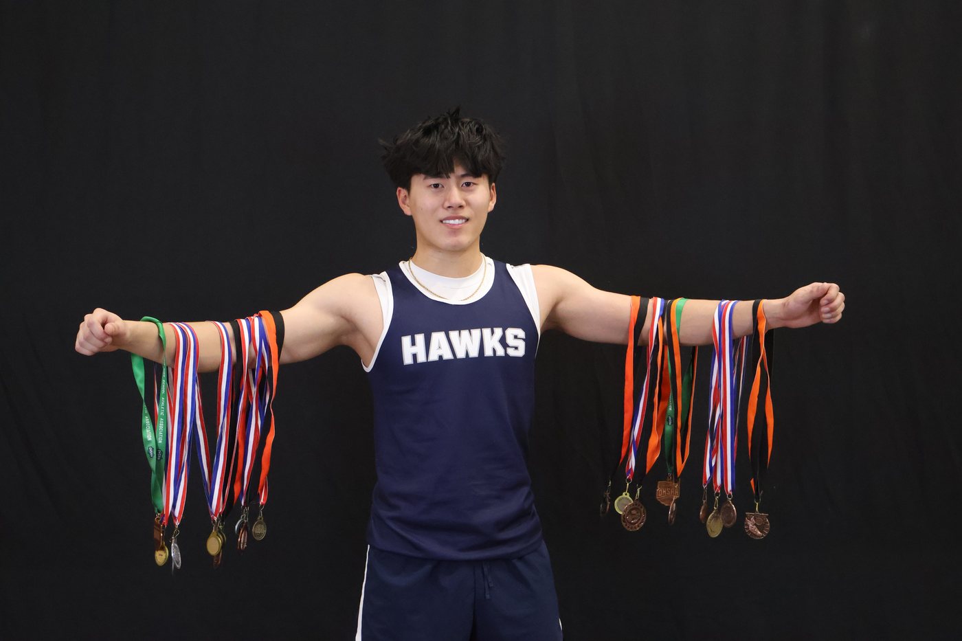 Joshua Wang in a Hawks track singlet with medals around his neck, arms outstretched.