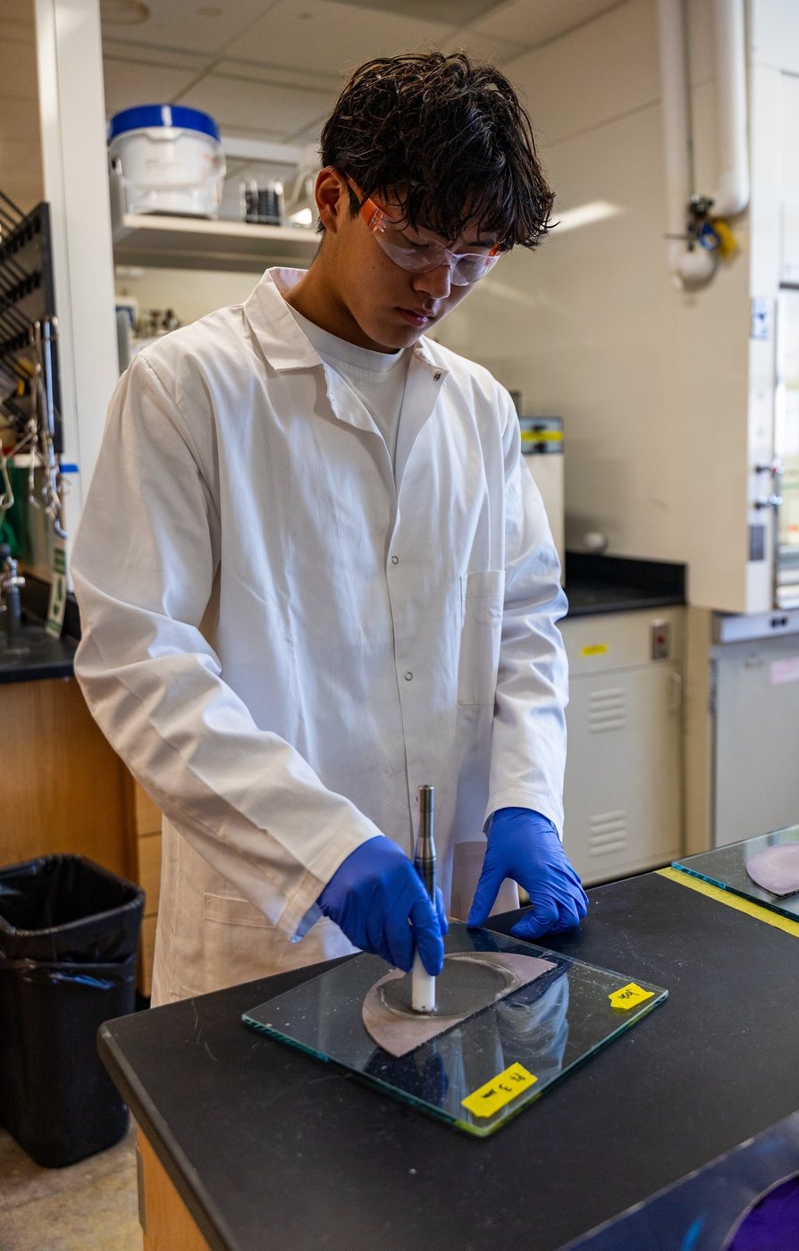 Joshua Wang in a white lab coat, safety glasses, and gloves working with a sample at a bench.