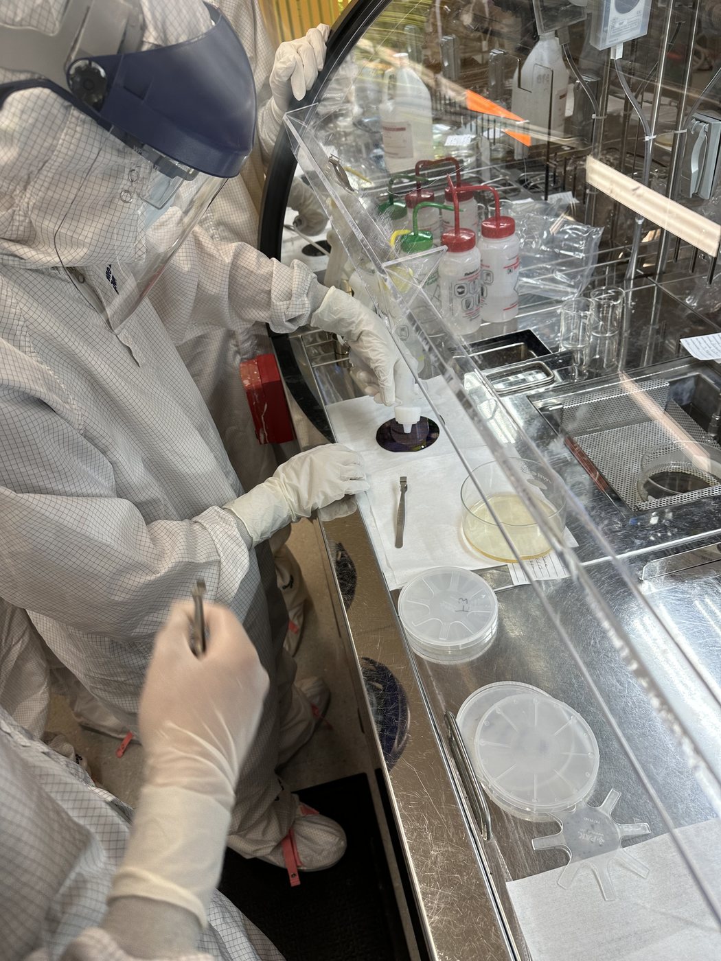 A researcher in protective gear and gloves reaching into a laboratory glovebox enclosure.